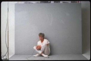Fotoshooting einer Performance- oder Schauspielgruppe : Ein Schauspieler in weißem Kostüm vor einem grauen Hintergrund sitzend