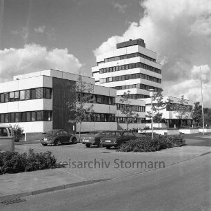 Rathaus: Blick von Südosten: davor Fahnenmast, Straßenlampe, Parkplatz mit Einfahrt und Autos, Grünstreifen mit jungen Bäumen: im Vordergrund Straße Reeshoop [später Manfred-Samusch-Straße]