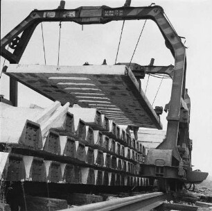 Rethwisch. Betonwerk der Deutschen Reichsbahn. Lagerplatz mit schienenverfahrbarem Portralkran beim Transport einer Schwellenladung mit Traversen