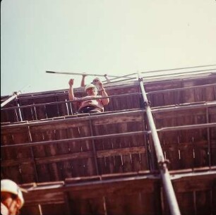 Zimmerer auf einem Gerüst beim vertikalen Weiterreichen einer Holzbohle
