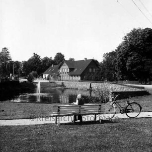 Dorfstraße: Dorfteich mit Springbrunnen: vorn Grünanlage: in der Mitte Bank mit Frau: daneben Fahrrad: hinten Häuser und Bäume