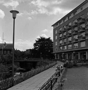 Grünanlage mit Fußweg, Jägerzaun und Straßenlampe: links Trave: rechts Wohnblock: hinten links Brücke an Hagenstraße: dahinter links Gebäude