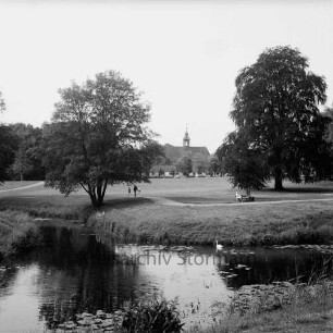 Schlosspark: äußerer Schlossgraben mit Seerosen, Schwan: Spaziergänger auf Fußweg, Frau auf Parkbank, Bäume: im Hintergrund Gottesbuden, Schlosskirche