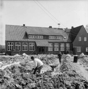 Volksschule: Königstraße: Ausbau zur Grundschule: Erweiterungsbau mit Anbau und Gymnastikhalle: vorn Arbeiter zwischen Erdhaufen beim Mauern der Grundmauern: daneben Schubkarre, Mörtelbottich und Mauersteine: dahinter Mann und Tonne: hinten Rückfront des Altbaus