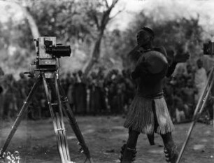 Tanz vor der Kamera : Nordrhodesien (heute: Samibia). Loangwatal (heute: Luangwa-Tal). Tänzer mit Beinrasseln, Handtrommel und angeklebtem Bart vor der Kamera, 11. Oktober 1931