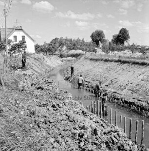 Barnitz: Flussregulierung: Bauabschnitt bei der Ölmühle: Ausbau der Uferböschung: vorn Mitte Arbeiter im Wasser beim der Holzstacks: links auf Böschung Zuschauer: dahinter Haus: hinten rechts Erdhaufen, Baubude und Baufahrzeug: im Hintergrund Bäume