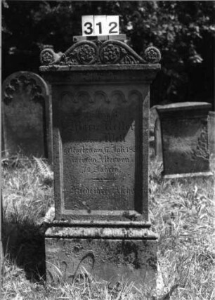 Grabstein 312 (Index: 100085232) Maße HxBxT: 123x55x43 cm Beschreibung: Stele, profilierter Sockel, mit Volutenaufsatz, Rundbogenfries, Schrift eingetieft: Grabstein 312