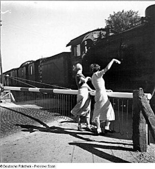 Zwei Mädchen an einem beschrankten Bahnübergang, einem vorbeifahrenden Personenzug zuwinkend