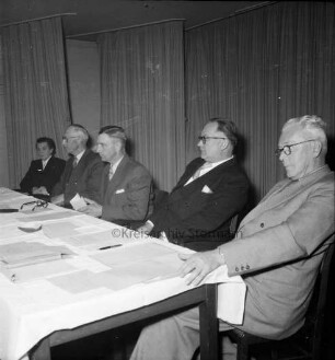 Keck, Erika: Kreispräsidentin: Kreisberufschule Bad Oldesloe: Kreistagssitzung am 18. Mai 1955: Verabschiedung: Präsidiumstisch mit dem neuen Kreispräsidenten Friedrich Hardt und den Stellvertretern Willy Esser und Paul Manthey sowie Landrat Wilhelm Siegel: hinten Vorhänge
