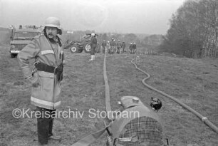 Großübung: Klein Wesenberg: Feuerwehrmann auf Weide vor Motorpumpe für Feuerwehrschläuche: im Hintergrund weitere Feuerwehrmänner mit Feuerwehrschläuchen neben Traktor und Einsatzwagen: rechts Wäldchen: im Hintergrund Bäume und einzelne Häuser