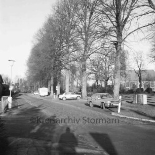 Kirchenweg: Kopfsteinpflaster: in der Mitte in Parkbuchten Pkws und Wohnwagen: dahinter Baumreihe des Dorfangers: hinten rechts Friedenskirche und Kirchhof: links Straßenlampe und Häuser