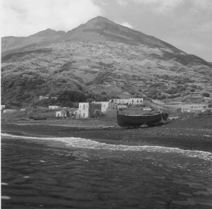Stromboli, Blick vom Schiff über den Strand auf Häuser des Ortes und die Insel