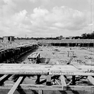 Freibad: Reeshoop: Nichtschwimmerbecken im Bau: Fundament fertig, Beckenwände mit Verschalung, Gerüst, Leitern: auf Beckensohle drei Arbeiter, Schalbretter, Gerüstbalken, Holzböcke, Arbeitsbühne: im Vordergrund Holzplanke,: hinten Bauzaun, Mann, links Holzschuppen: im Hintergrund Häuser Am Tiergarten