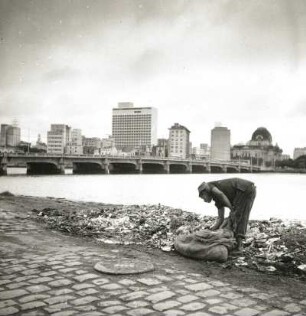 Recife, Brasilien. Mann beim Durchsuchen einer Müllhalde am Flußufer. Blick über den Rio Capibaribe gegen das Stadtzentrum