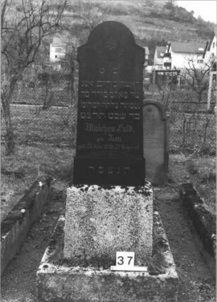 Grabstein 37 (Index: 99805059) Material: geschliffener Granit Zustand: gut Maße HxBxT: 149x53x28 cm Beschreibung: Stele auf Sockel mit eingezogenem Rundbogen Symbolik: eingemeißelter Stern mit Kranz im Rundbogen: Grabstein 37