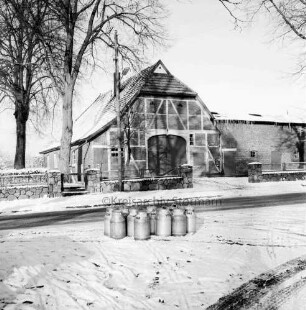 Dorfstraße mit Bauerngehöft: davor Vorgarten mit Feldsteinmauer und Bäume: vorn Milchkannen am Straßenrand: Winter