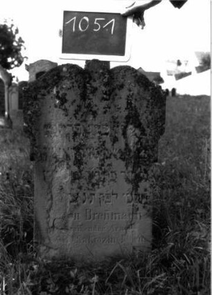 Grabstein 1051 (Index: 100104069) Material: Sandstein Zustand: stark verwittert und bewachsen Maße HxBxT: 76x47x13 cm Beschreibung: Stele mit seitlich auskragendem, geschweiftem Abschluss, in der Mitte eingezogen und spitz erhöht: Grabstein 1051