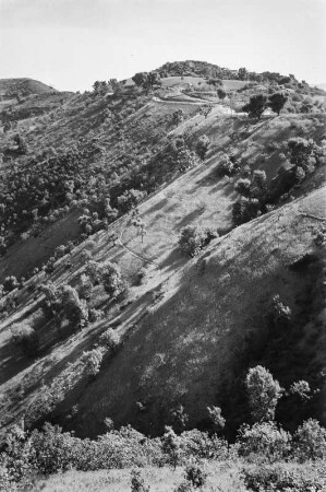 Dorf und Landschaftsblick : Blick auf das Dorf At Hicem, das auf einer Bergkuppe gelegen ist. An den Berghängen lassen sich Gerste- und Weizenfelder sowie Feigenbäume, Macchiebüsche, Eichen und Eschen erkennen.