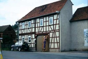 Haus mit Torfahrt, Fachwerk, am Hoftor arbeitet ein Mann auf einem Stuhl am Holz