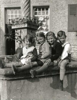 Vier Kinder, auf dem Rand eines Brunnenbeckens sitzend
