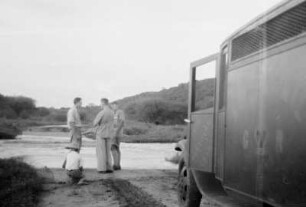 Auf der Landstraße : Blick auf eine Gruppe von Expeditionsteilnehmern und ein Automobil an einem Fluss