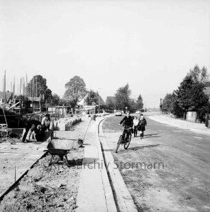Am Neuen Teich: Befestigen von Fußgängerweg und Radweg: Arbeiter beim Verlegen von Betonplatten: Stapel von Betonplatten, Schubkarre, Blechtonne: links Haus in Bau, mit Gerüst: Straße mit neuer Asphaltdecke, Junge auf Fahrrad, Kinder, hinten Auto: hinten rechts und links Einzelhäuser