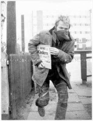 Junger Mann, halb vermummt, rennt mit einem Schild unterm Arm (Sonderthema: "Ich will ja nur dein Bestes." Eltern, Lehrer, Vorgesetzte)