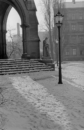 Blick auf einen winterlichen Kirchhof [vermutlich Peterskirche] und die umgebende Bebauung, im Zentrum eine sakrale Sandsteinplastik