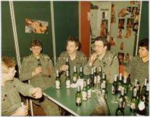 Junge Wehrdienstleistende sitzen um einen Tisch herum und trinken Bier, im Hintergrund Poster von nackten Frauen (Altersgruppe 18-21)