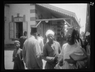 Zagazig (Ägypten): "Effendi [=Herr], Zeitungsverkäufer und Lastträger in typischen Trachten" vor dem Gebäude der Poststation (oder dem Bahnhof (?)) mit farblich abgesetzten Simsen und Wandkanten, Uhr und überdachtem Vorplatz
