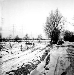 Wirtschaftsweg: vorn Mitte Graben: rechts und links Weiden und Felder mit landwirtschaftlichem Gerät: hinten rechts Bäume: hinten links Strommasten der Überlandleitung: Winter