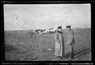 Nicht identifizierbare Ortschaft in Nordfrankreich oder Belgien: 2 Offiziere im Gespräche auf einem weiten Feld, im Hintergrund ein Doppeldecker und ein Kampfflugzeug mit Tatzenkreuz