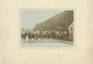 13 Offiziere in Uniform mit Mütze auf Ausritt, im Hintergrund eine Ortschaft