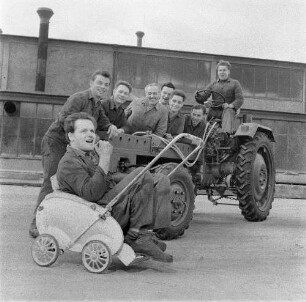 Schönebeck. VEB Traktorenwerk Schönebeck. Produktionsbereich Geräteträger RS 09. Gruppe Kraftfahrzeugschlosser an und auf einem Fahrzeug auf dem Werkgelände, davor ein Schlosser in einem Kinderwagen sitzend