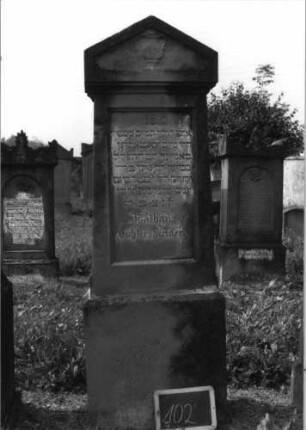 Grabstein 102 (Index: 99937109) Material: Sandstein Zustand: verwittert Maße HxBxT: 173x48x23 cm Beschreibung: konische Stele, Sockel, Giebel, rechteckiges Schriftfeld eingetieft Symbolik: Levitenkanne: Grabstein 102