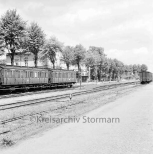 Bahnhofsgelände: Gleisanlagen, Bahnsteige, beschrankter Gleisübergang: Personenzug mit Einzelabteilen, Güterzug: hinten links Bahnhofsgebäude, Wartehäuschen, Personen, Bäume, rechts Nebengebäude
