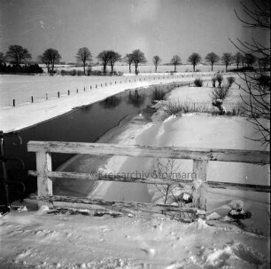 Trave: vorn Brücke der Landstraße Richtung Travenhof mit Holzgeländer: Blick ins Travetal mit Wiesen, Baumreihen und Knicks: Winter