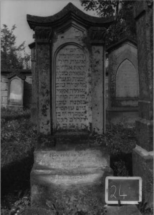 Grabstein 24 (Index: 99801893) Material: Sandstein Zustand: verwittert Maße HxBxT: 144x47x22 cm Beschreibung: ädikulaartige Stele mit Pilastern, getreppter Sockel, geschwungener giebelartiger Abschluß mit Verdachung, eingetietes Schriftfeld mit Rundbogen Besonderheit: seltene Form: Grabstein 24