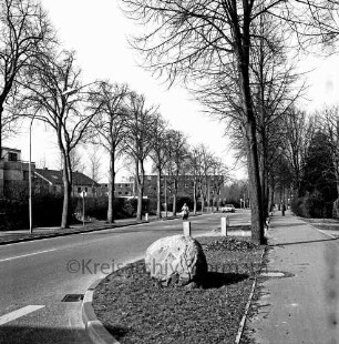 Sieker Landstraße: rechts Fußgängerweg: darauf hinten Mann: vorn Grünfläche mit Findling: dahinter Baumreihe: hinten Mitte Mann auf Motorroller und Pkw: dahinter weitere Baumreihe und Gebäude