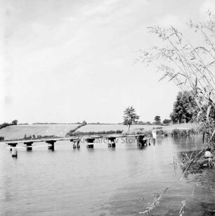 Moorteich mit Schulschwimmsteg: rechts Schilf: hinten rechts Wanderweg und Bäume: im Hintergrund Knicklandschaft