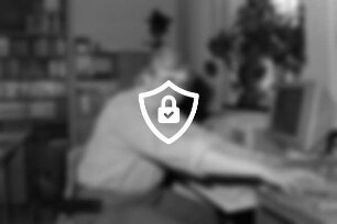 Mitarbeiterin [Name maskiert] sitzt vor einem Schreibtisch mit Computer und greift nach [Name maskiert] Filmdosen: im Hintergrund Regale