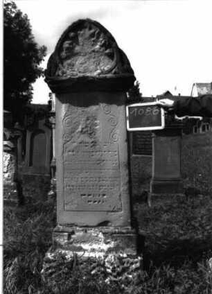 Grabstein 1086 (Index: 100104135) Material: Sandstein Zustand: stark verwittert mit fast vollständigem Textverlust Maße HxBxT: 200x49x23 cm Beschreibung: Stele auf getrepptem Sockel, getreppter Aufsatz mit hohem Bogenabschluss, ausgefüllt mit Akanthus, Mohnkapseln und Voluten (verwittert), Schriftfeld durch eingeritzte Linie gerahmt, die oben bogenartig in Voluten ausläuft, unterer Sockel felsartiggearbeitet Symbolik: Akanthus, Mohnkapseln, Voluten Besonderheit: kulturhistorisch wertvoll / hist.-wiss. Gründe (Vorbeter): Grabstein 1086
