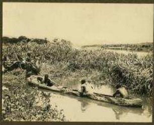 Einbaum mit Afrikanern in Ufernähe bei wartendem Askari mit Trägern