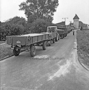 Wiesenstraße: Schlange von landwirtschaftlichen Fuhrwerken mit beladenen Anhängern: hinten rechts Silo der Raiffeisengenossenschaft: davor Mann auf Straße: links Büsche und Bäume