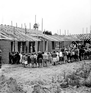 Volksschule: Neubau: Rohbau: Richtfest: vorn Sand- und Erdhaufen und Gestrüpp: dahinter Schülerhalbkreis: rechts dahinter Funktionsträger: hinten Pavillonbauten mit Richtkrone und Arbeitern auf Dachsparren