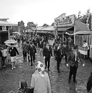 Schützenfest: Veranstalter Bürgerschützengilde von 1627 e.V. Bad Oldesloe: Umzug: Festplatz: Sehmsdorfer Straße: Einmarsch der Schützenvereine mit Fahnen, Schützen gehen über Pfützen: Jahrmarktstände, links "Schießhalle", davor Soldaten: Besucher mit Regenschirmen, schlammiger Boden, 25. Juli 1965