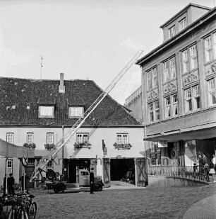 Hude: Feuerwehrgerätehaus "Blaues Haus" mit geöffneten Toren: links Feuerwehrfahrzeug mit ausgefahrener Drehleiter: daran Feuerwehrmänner: rechts Wohn- und Geschäftshaus Gebrüder Petersen und Zuschauer