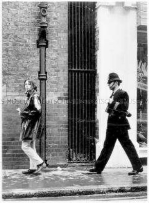 Junge Frau auf dem Gehweg, hinter ihr ein britischer Polizist