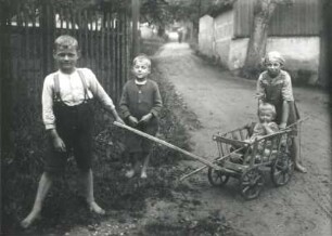 Zwei Jungen und eine Mädchen mit Kleinkind im Leiterwagen