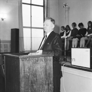 Theodor-Mommsen-Schule: Aula: Theodor-Mommsen-Fest: vorn Rednerpult mit Redner: dahinter auf der Bühne sitzende Schülerinnen und Mikrofon: 7. Dezember 1968